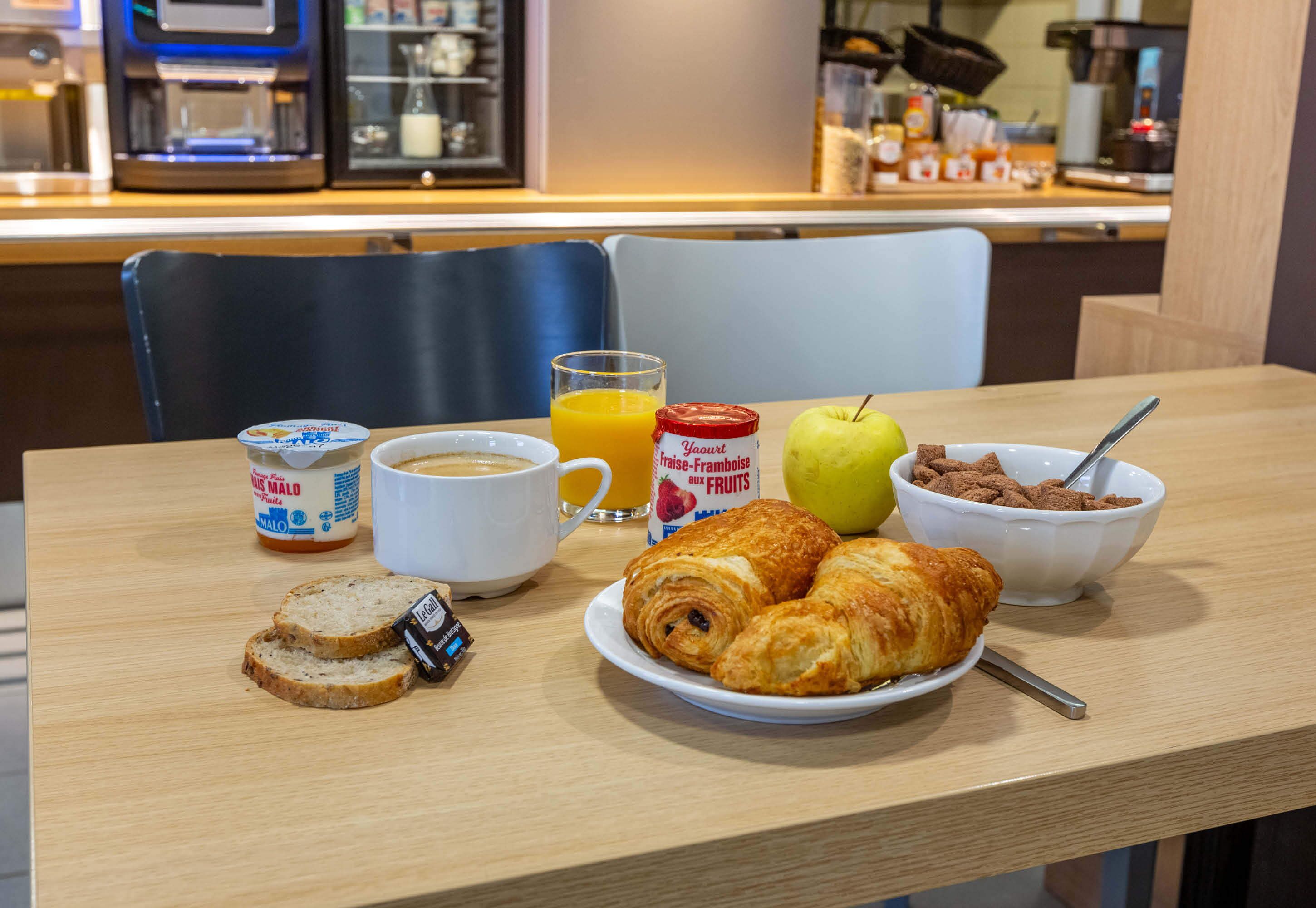A breakfast table with croissants, a bowl of cereal, a glass of orange juice, a cup of coffee, and a jar of yogurt.