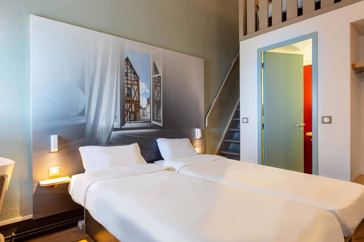 A hotel room with a large bed, a painting of a building on the wall, a nightstand with a lamp, and a staircase leading to an upper level.
