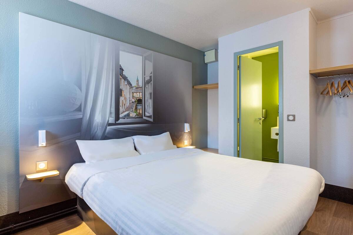 A hotel room with a large bed, a nightstand with a lamp, a wall-mounted artwork, a green door, and a coat rack.