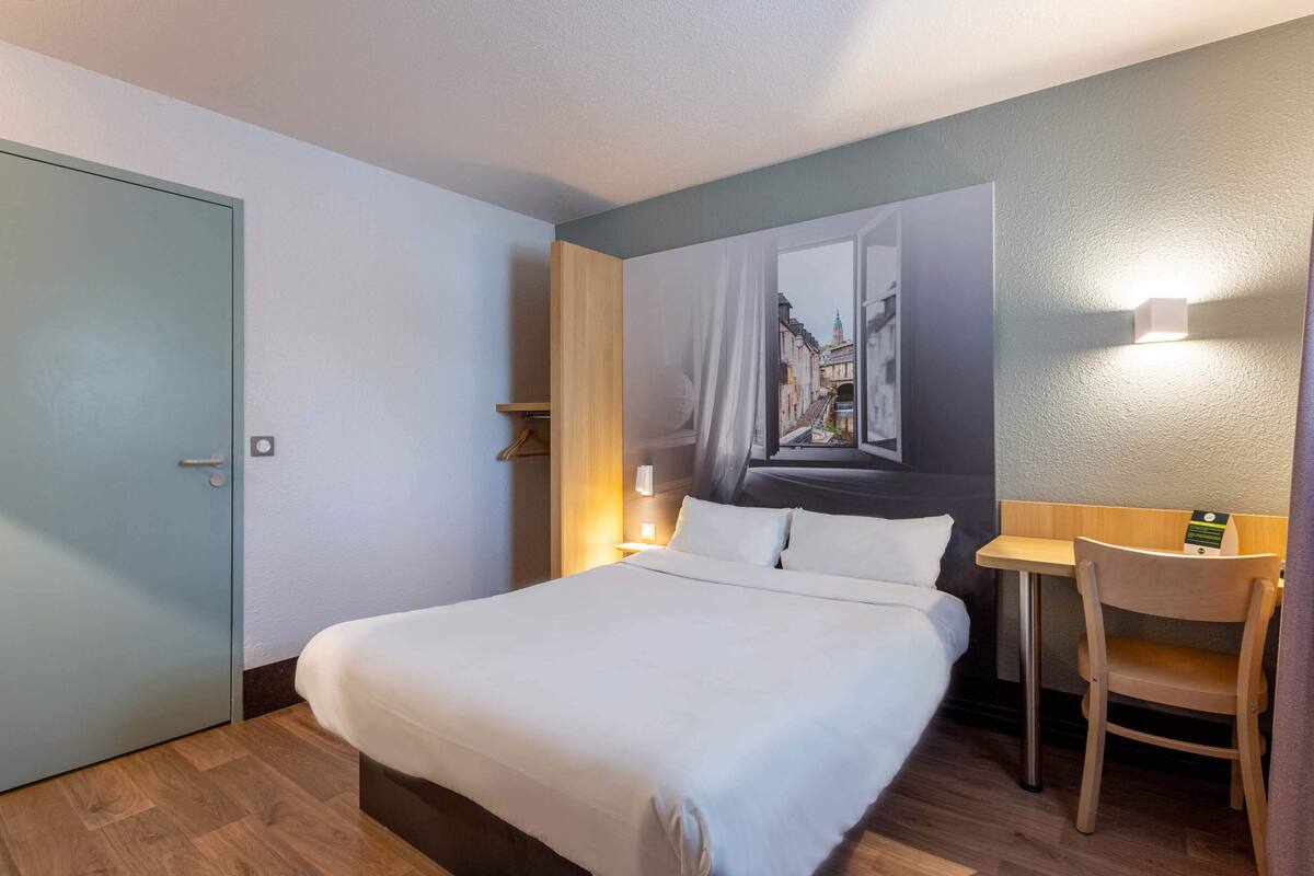 A hotel room with a bed, a desk, a chair, a nightstand, and a wall-mounted lamp.