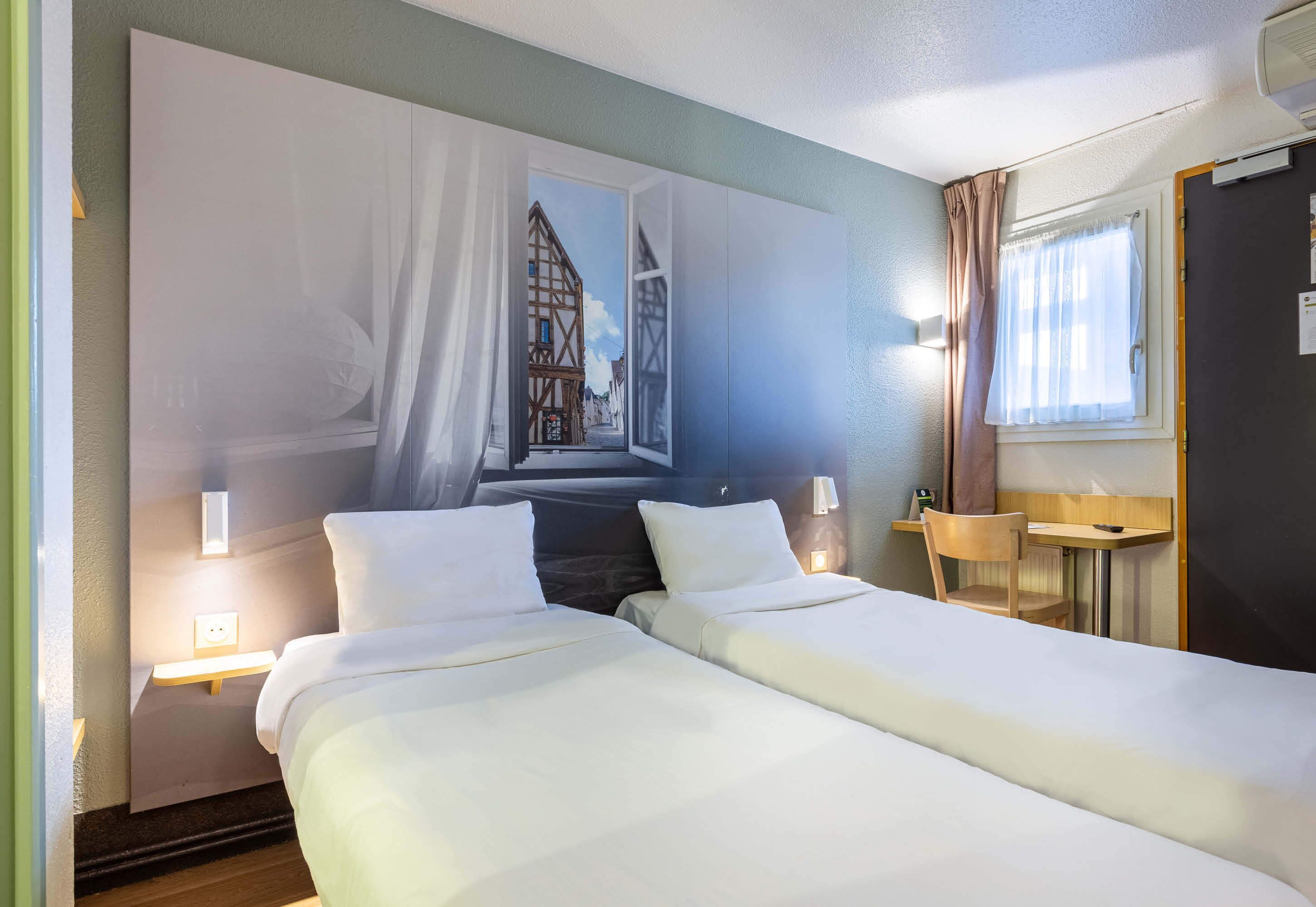 A hotel room with two beds, a wall mural, a bedside table with a lamp, a window with curtains, and a small wooden table.