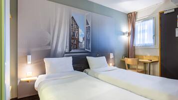 A hotel room with two beds, a wall mural, a bedside table with a lamp, a window with curtains, and a small wooden table.