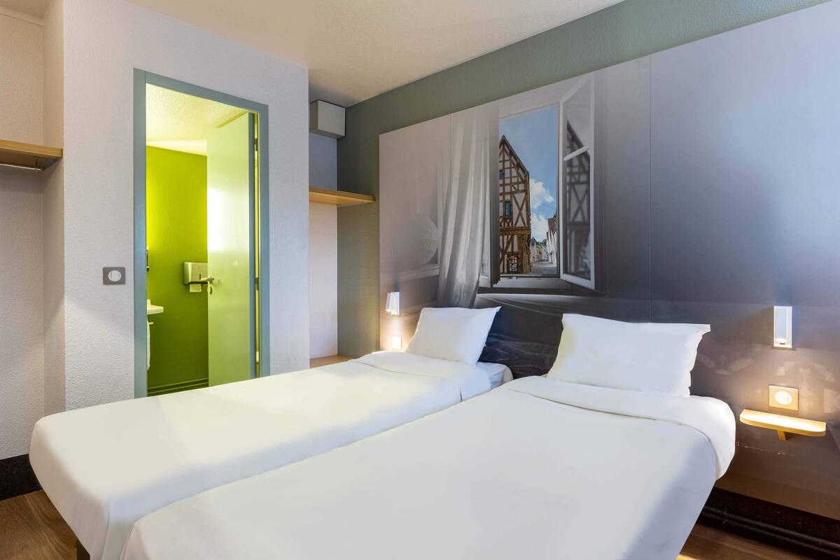 A hotel room with two beds, a green door, a painting of a building, and bedside lamps.