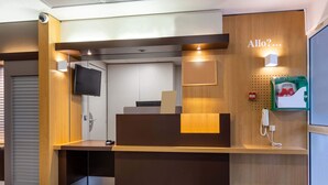 A reception desk with a flat-screen TV, a telephone, and a green waste bin. - B&B HOTEL CHARTRES Le Forum (1) (Le Coudray)