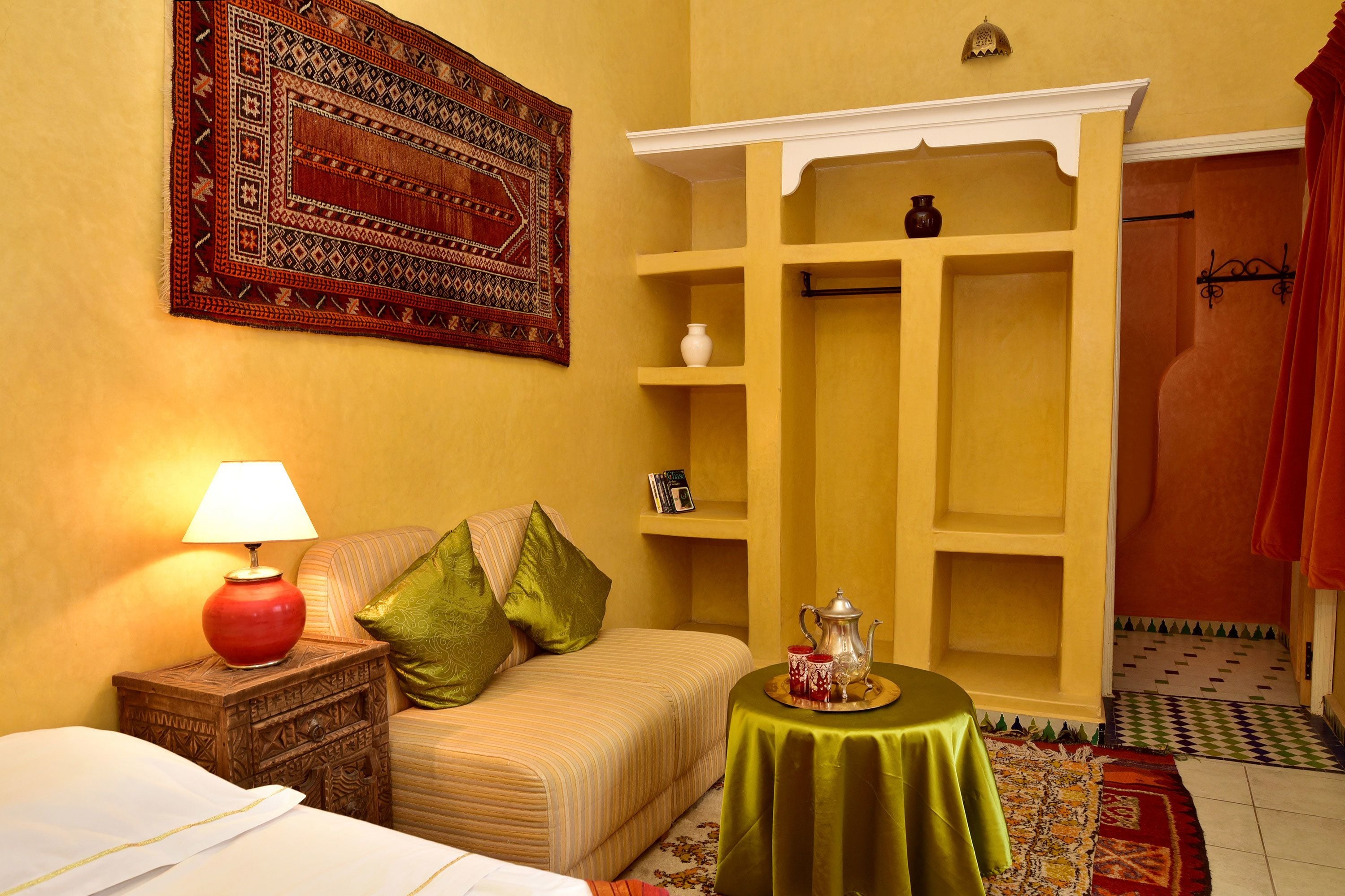 A room with a bed, a nightstand with a lamp, a green table with a tray, a shelf with a vase, and a patterned rug.