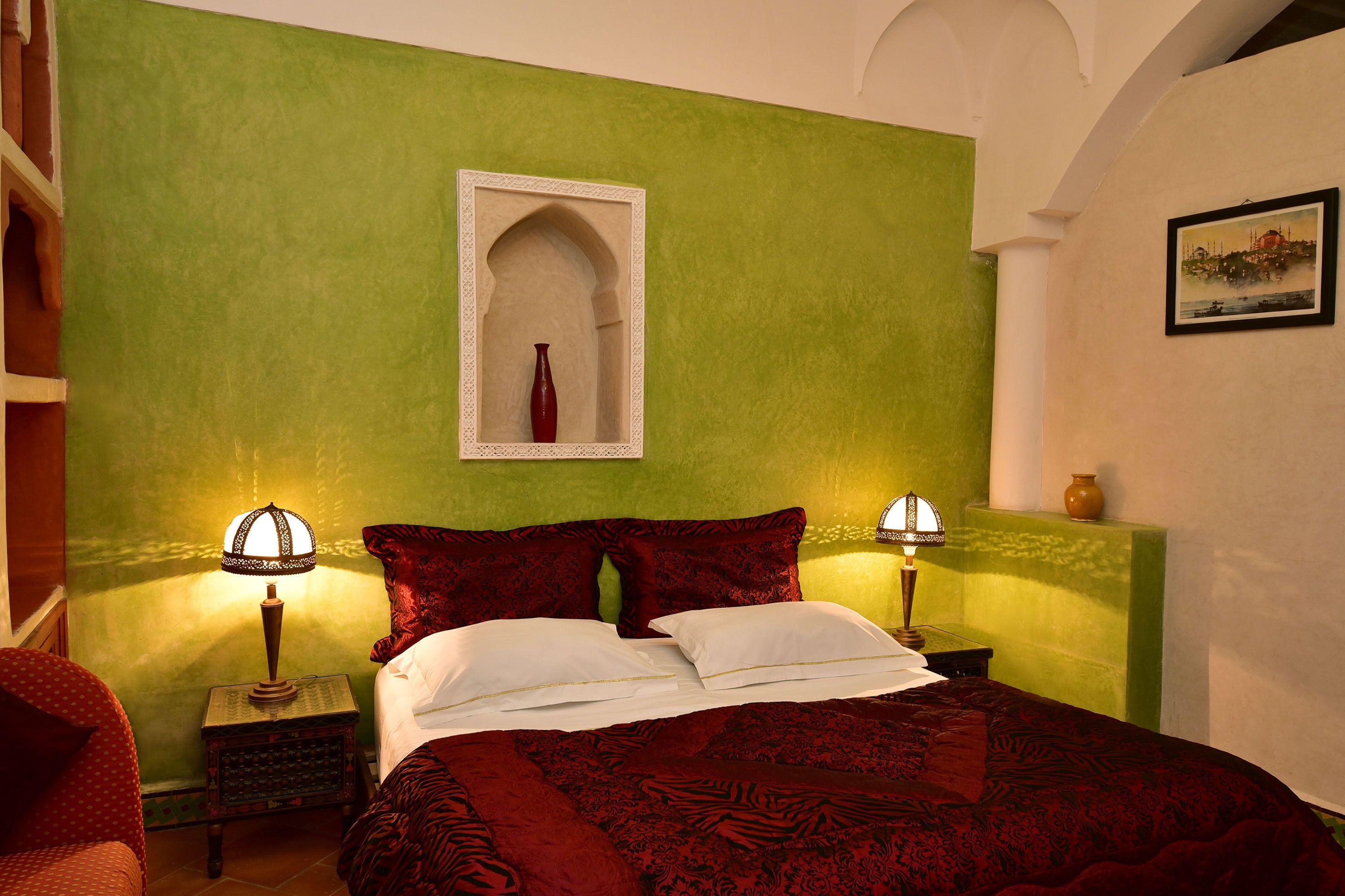 A bed with red bedding, two bedside lamps, a small table, and a wall-mounted mirror.