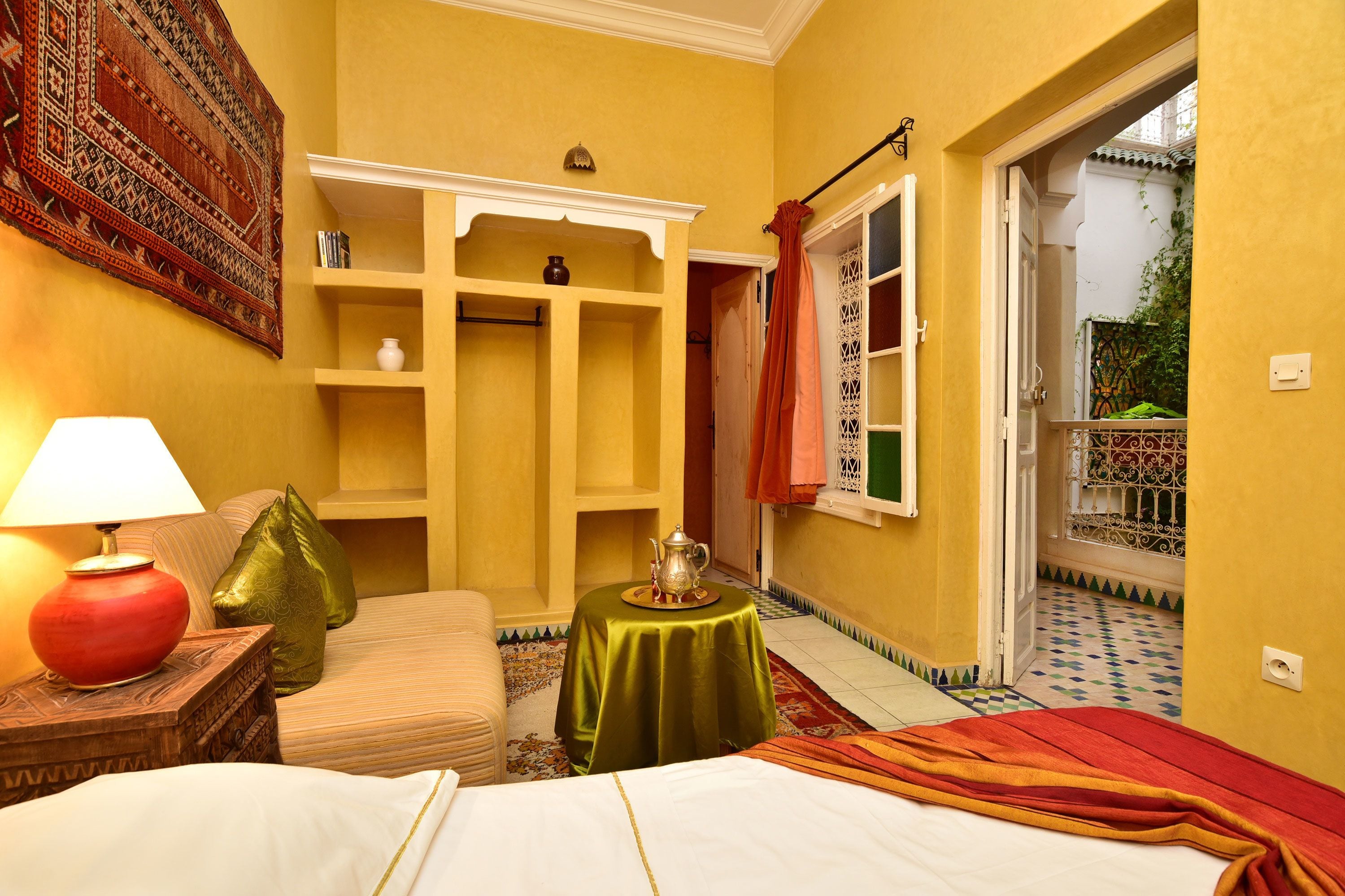 A hotel room with a bed, a small table with a teapot, a sofa, a bookshelf, and a view of the outside balcony.