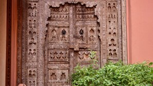 Una puerta de madera tallada tradicional con diseños intrincados, una planta en maceta al frente y un suelo de baldosas con motivos geométricos.