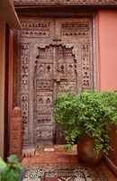 Una puerta de madera tallada tradicional con diseños intrincados, una planta en maceta al frente y un suelo de baldosas con motivos geométricos.