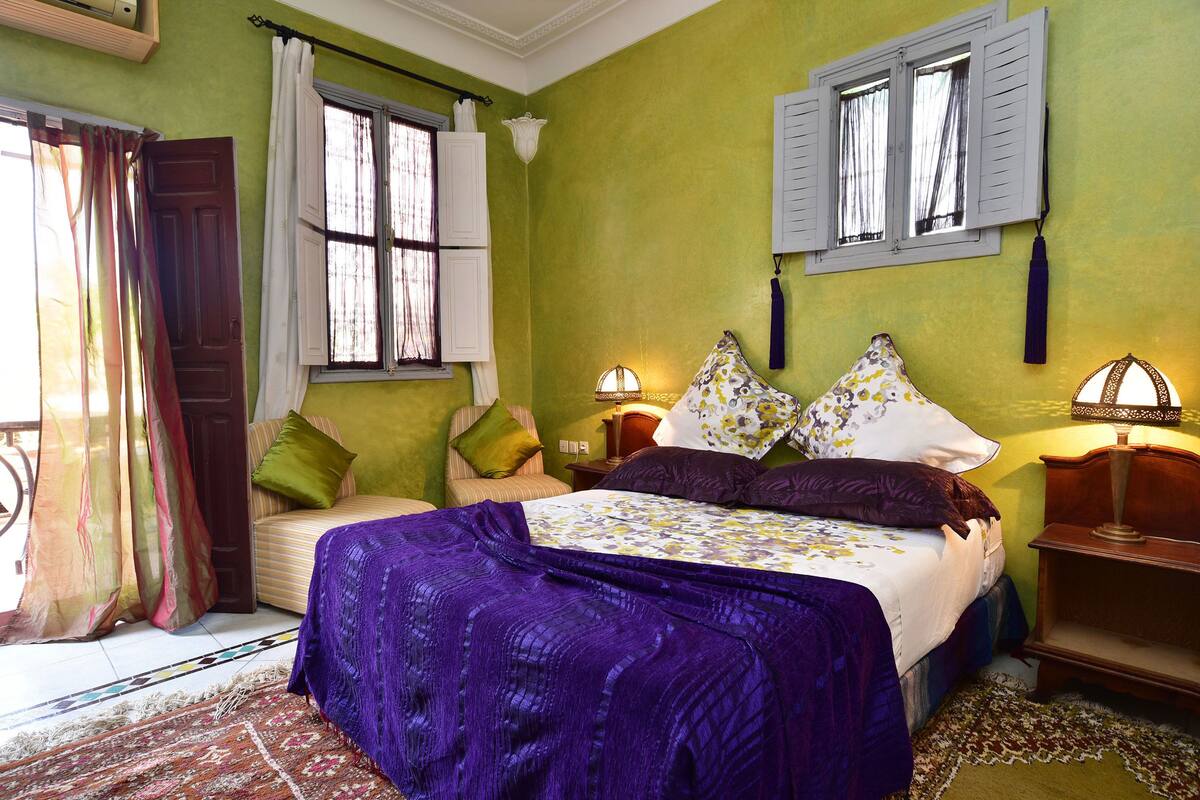 A room with a bed, two bedside tables, a window with shutters, and a patterned rug.