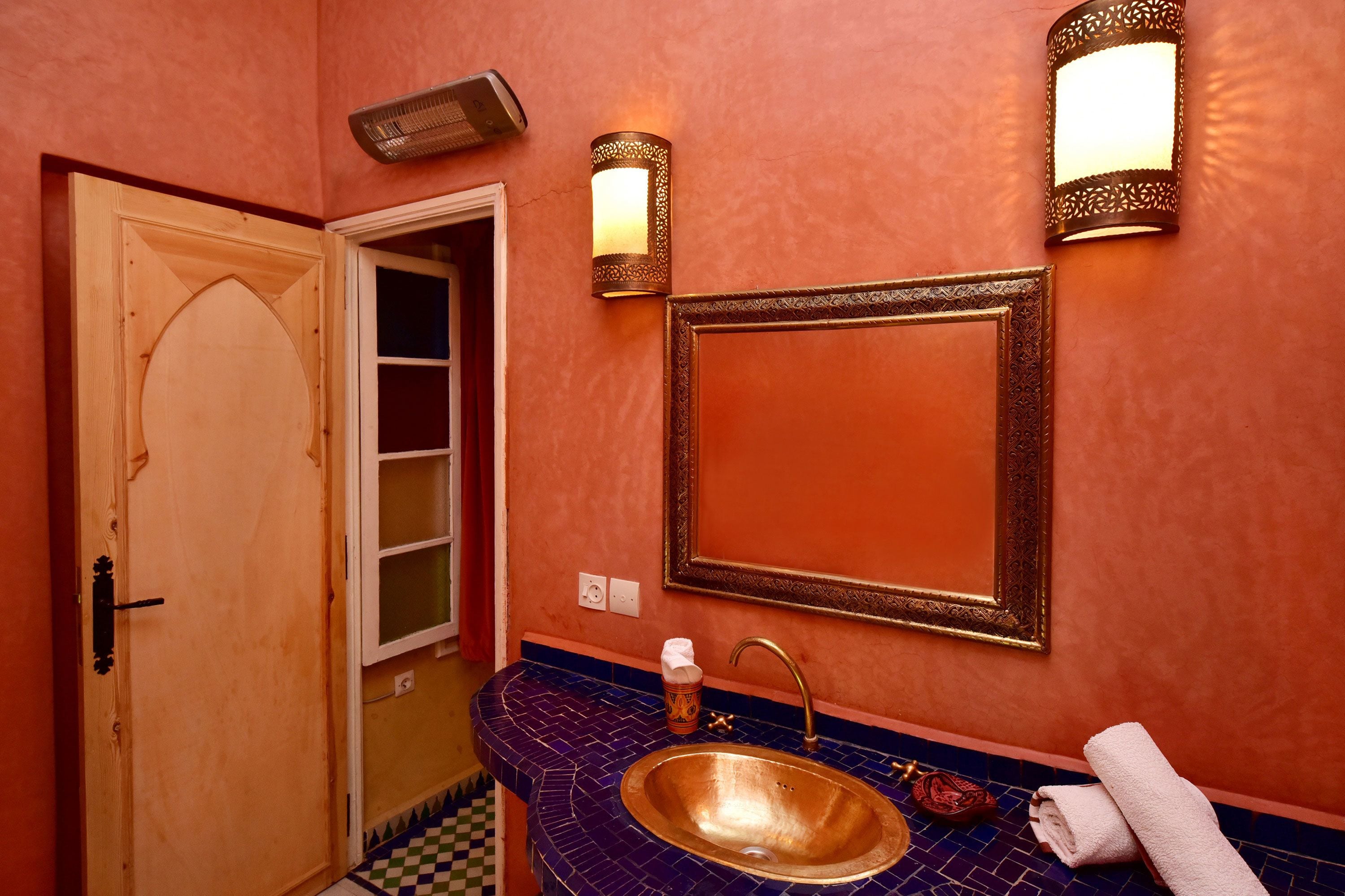 Un bany amb una porta de fusta, un lavabo amb taulell de rajola blava i dues làmpades penjades a la paret.