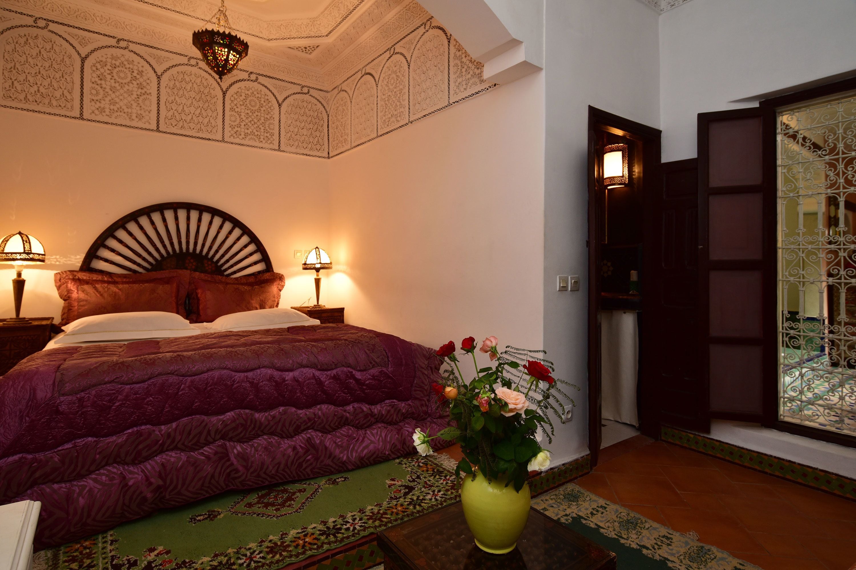 A bedroom with a large bed, two bedside lamps, a decorative ceiling, and a view through an open door.