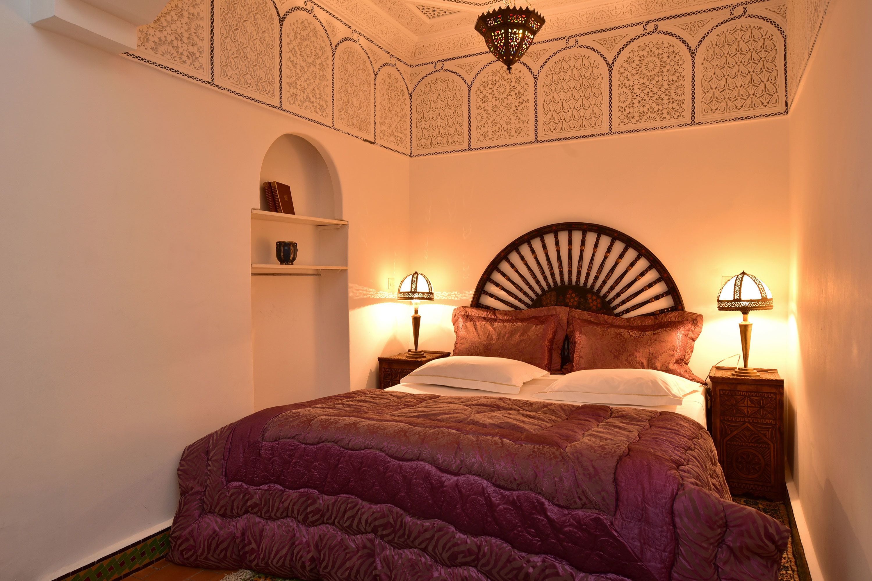 A bedroom with a large bed, two bedside lamps, a decorative ceiling, and a small shelf.