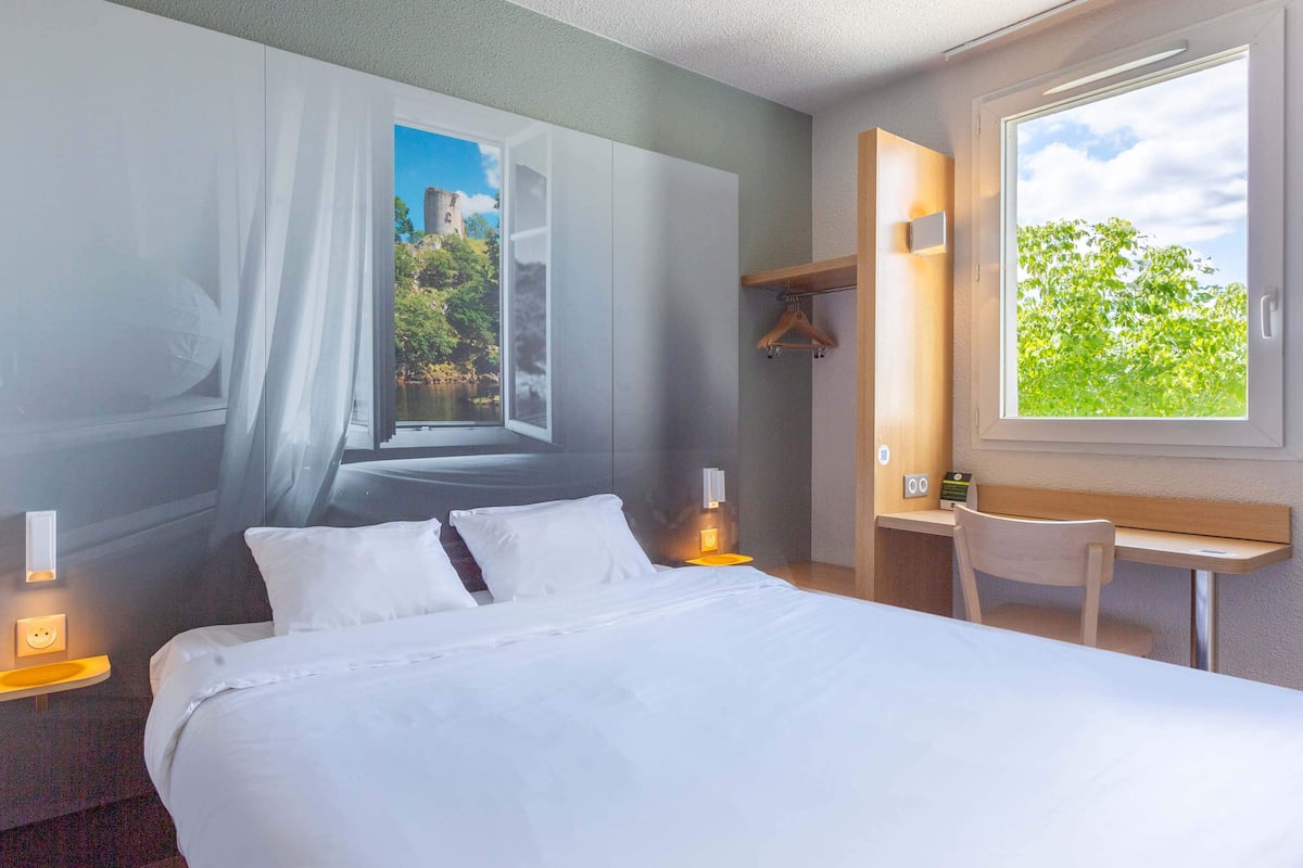 Ein Hotelzimmer mit Bett, Nachttisch, Lampe, Stuhl und einem Fenster mit Blick auf Grünflächen und ein Gebäude.