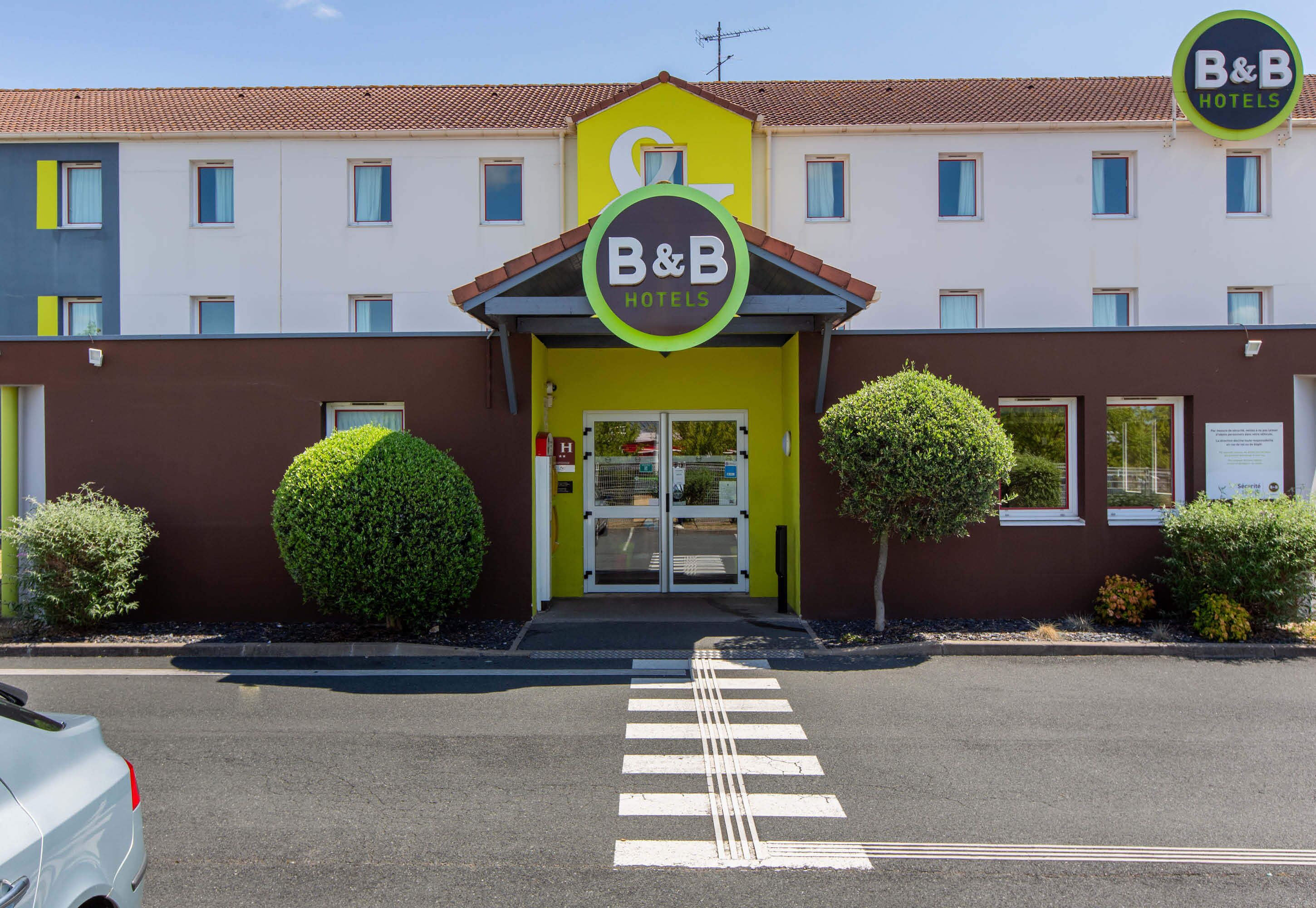 Un hôtel de type « bed and breakfast » avec une façade jaune, deux arbustes ronds et un ciel dégagé.