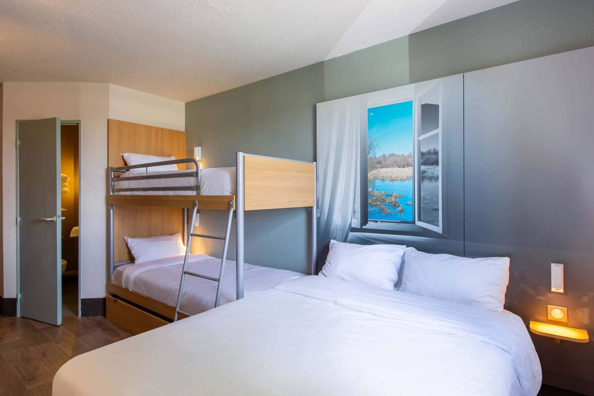 Ein Hotelzimmer mit einem Etagenbett, einem Einzelbett, einem Landschaftsbild und einem Fenster mit Blick auf ein Gewässer.