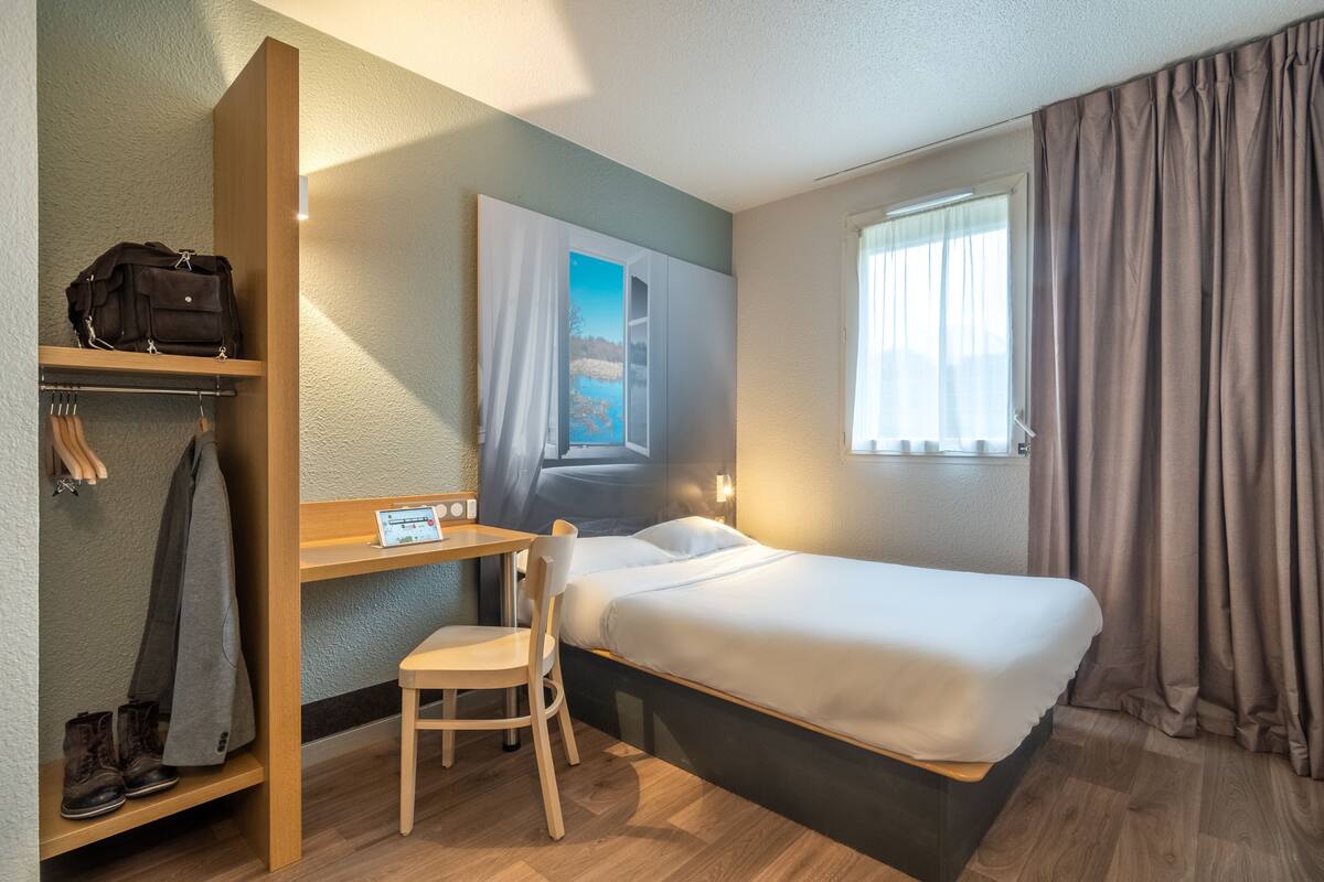 Ein Hotelzimmer mit einem Bett, einem Schreibtisch mit Stuhl, einem Kleiderständer und einem Fenster mit Vorhängen.