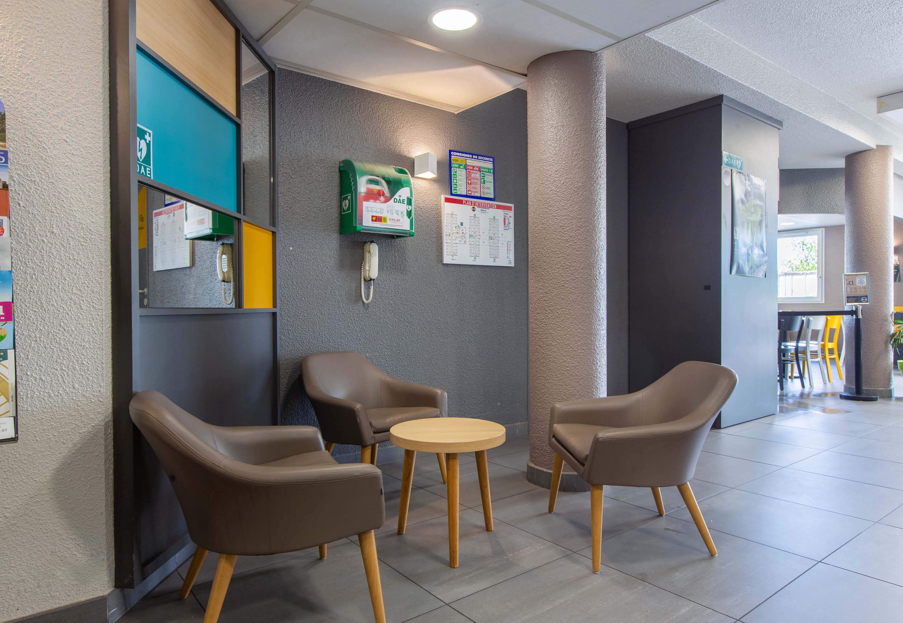 A hotel lobby with a reception desk, a green emergency phone, informational posters, and a seating area with two chairs and a small round table.