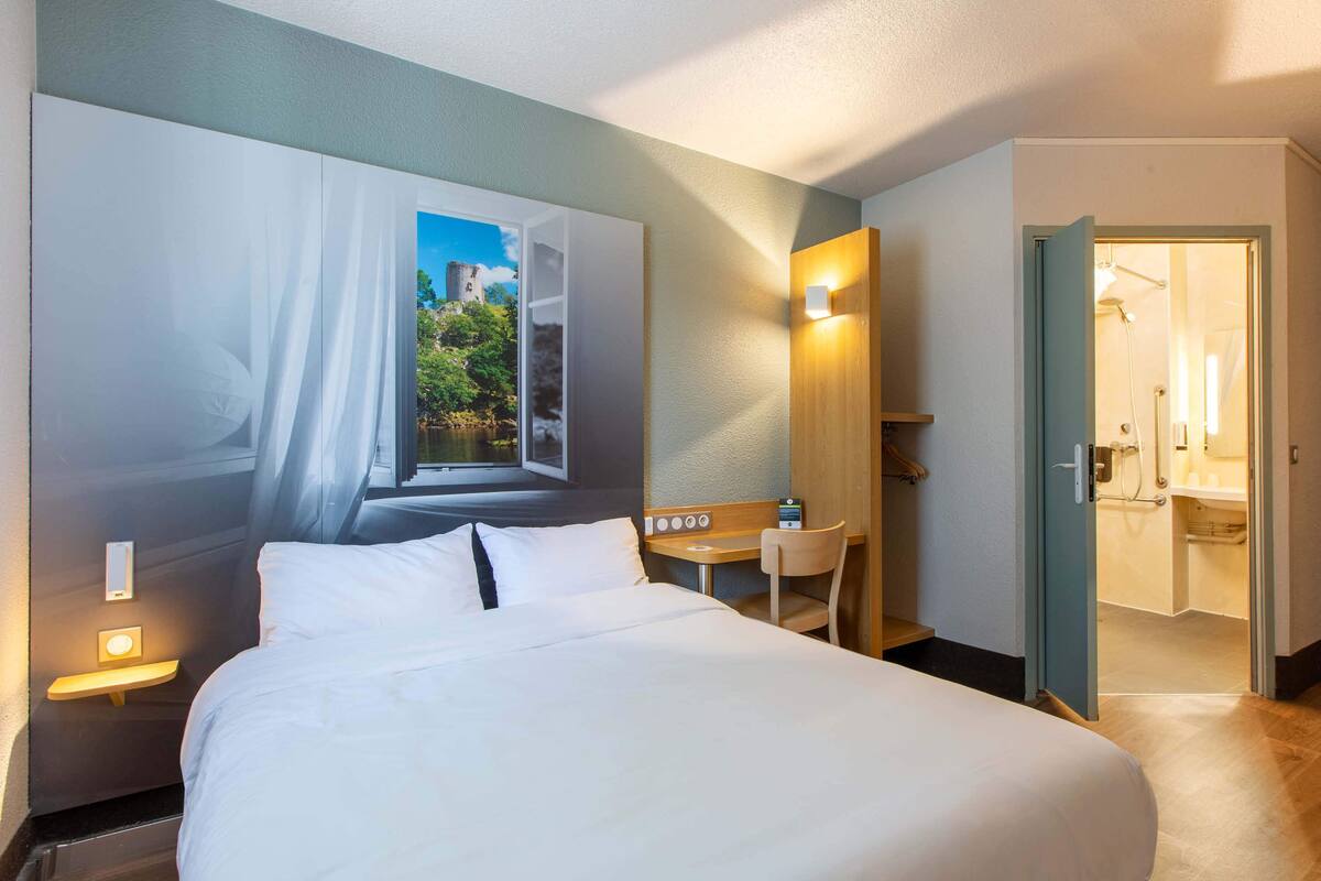 Ein Hotelzimmer mit Bett, Schreibtisch, Stuhl und Blick auf eine Stadtlandschaft durch ein Fenster.