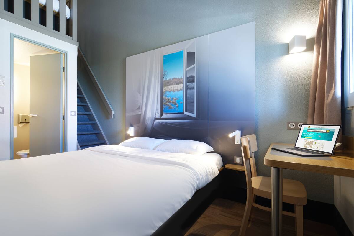 A hotel room with a bed, a desk with a laptop, a chair, a window with a view of a landscape, and a staircase.