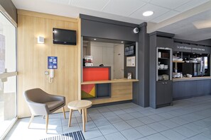 Eine moderne Hotelhalle mit einer Rezeption, einer Sitzecke mit Stuhl und kleinem Tisch sowie einer durch eine Glastrennwand sichtbaren Küchenzeile.