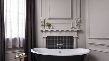 Une salle de bain avec une baignoire sur pieds, une petite étagère avec des objets décoratifs et une fenêtre avec des rideaux.