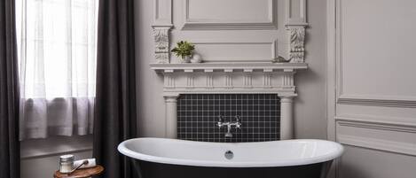 A bathroom with a freestanding bathtub, a small shelf with decorative items, and a window with curtains.