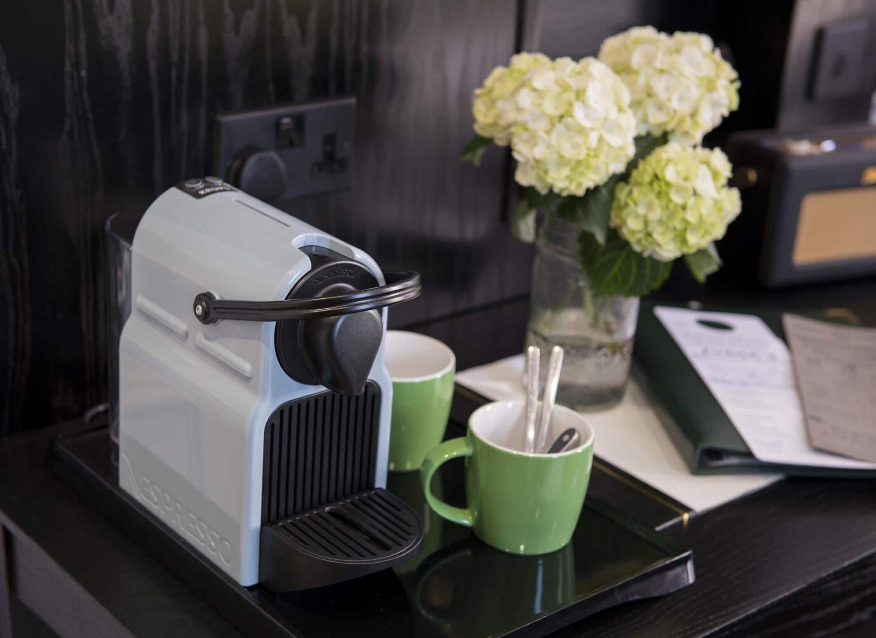 A coffee maker on a black tray with a green mug and a vase of white flowers.