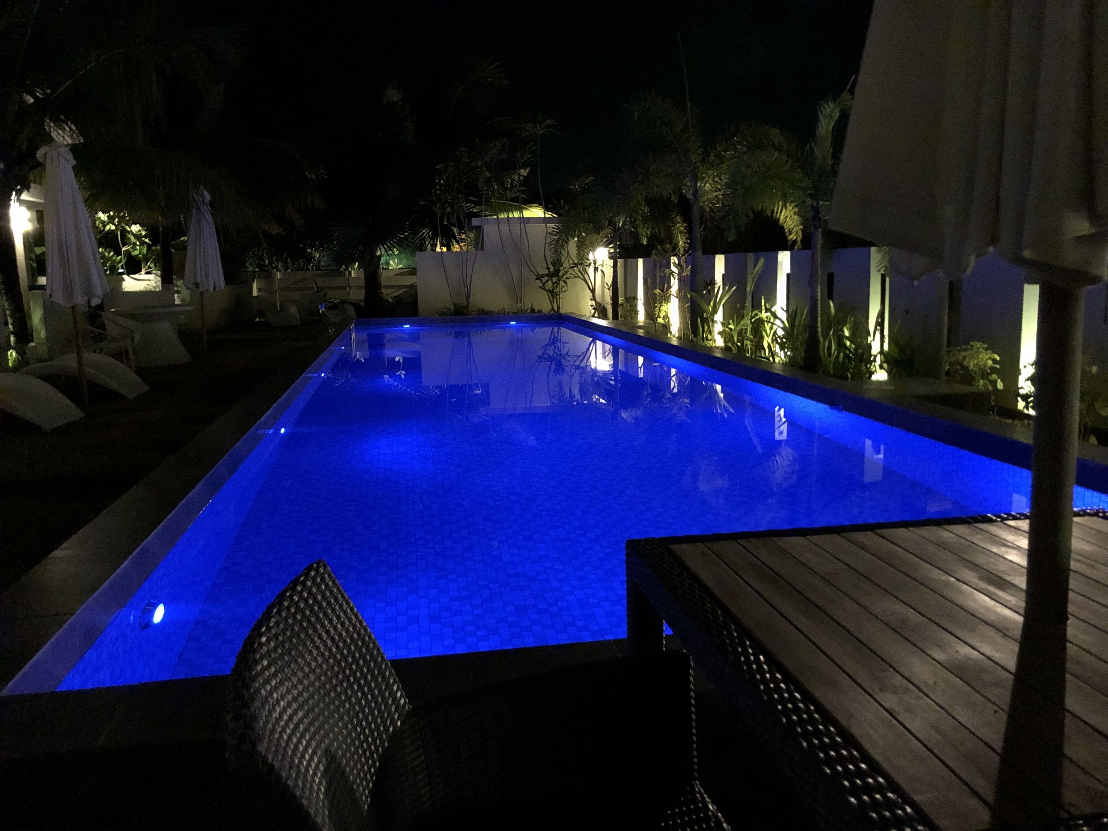 Piscina retangular com tonalidade azul, rodeada por espreguiçadeiras e guarda-sóis, e um deck de madeira.