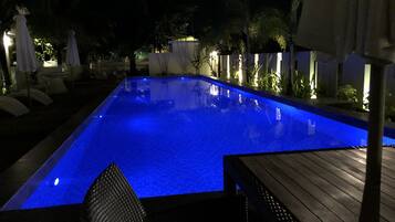 Piscina retangular com tonalidade azul, rodeada por espreguiçadeiras e guarda-sóis, e um deck de madeira.