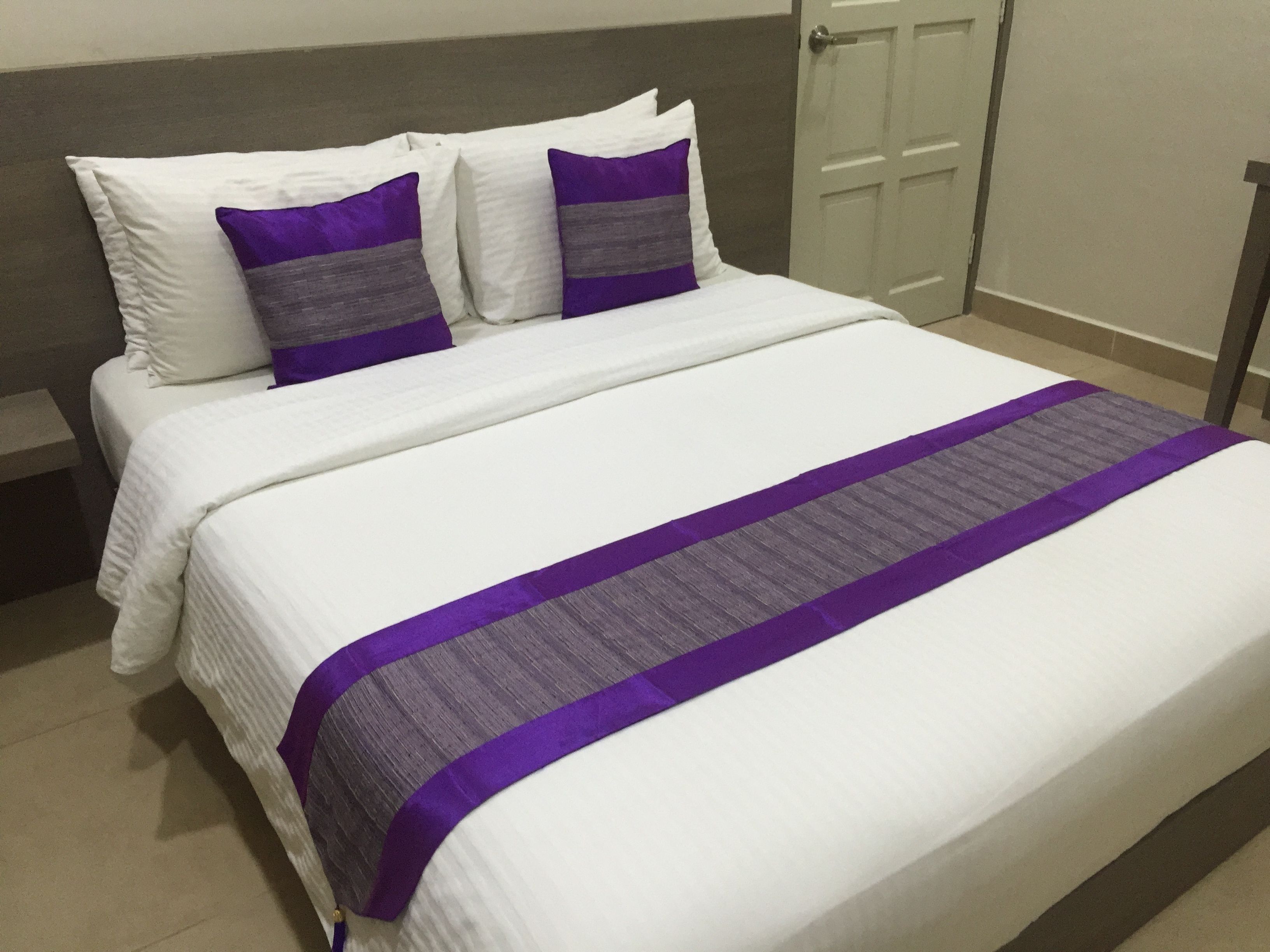 Uma cama bem arrumada com roupa de cama branca e detalhes em roxo, uma porta fechada ao fundo e piso de azulejos.