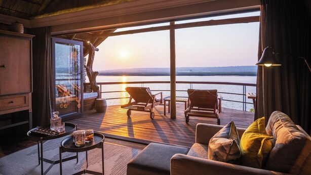 A cozy room with a large window overlooking a body of water at sunset. There are two wooden chairs and a sofa with cushions. - Chobe Water Villas (Kasika)