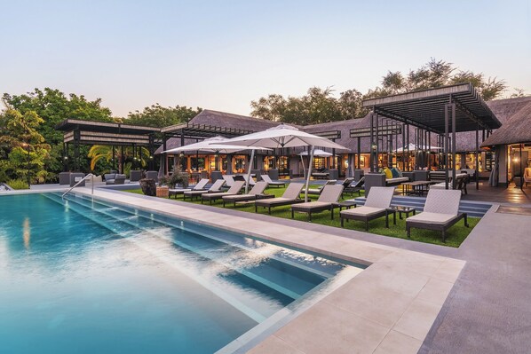 A poolside area with lounge chairs and umbrellas, a pool, and a building with a thatched roof.