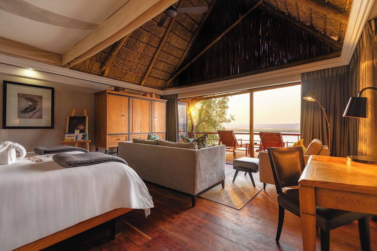 A hotel room with a bed, a sofa, a dining table, a chair, a desk, a lamp, a painting, and a view of the outdoors.