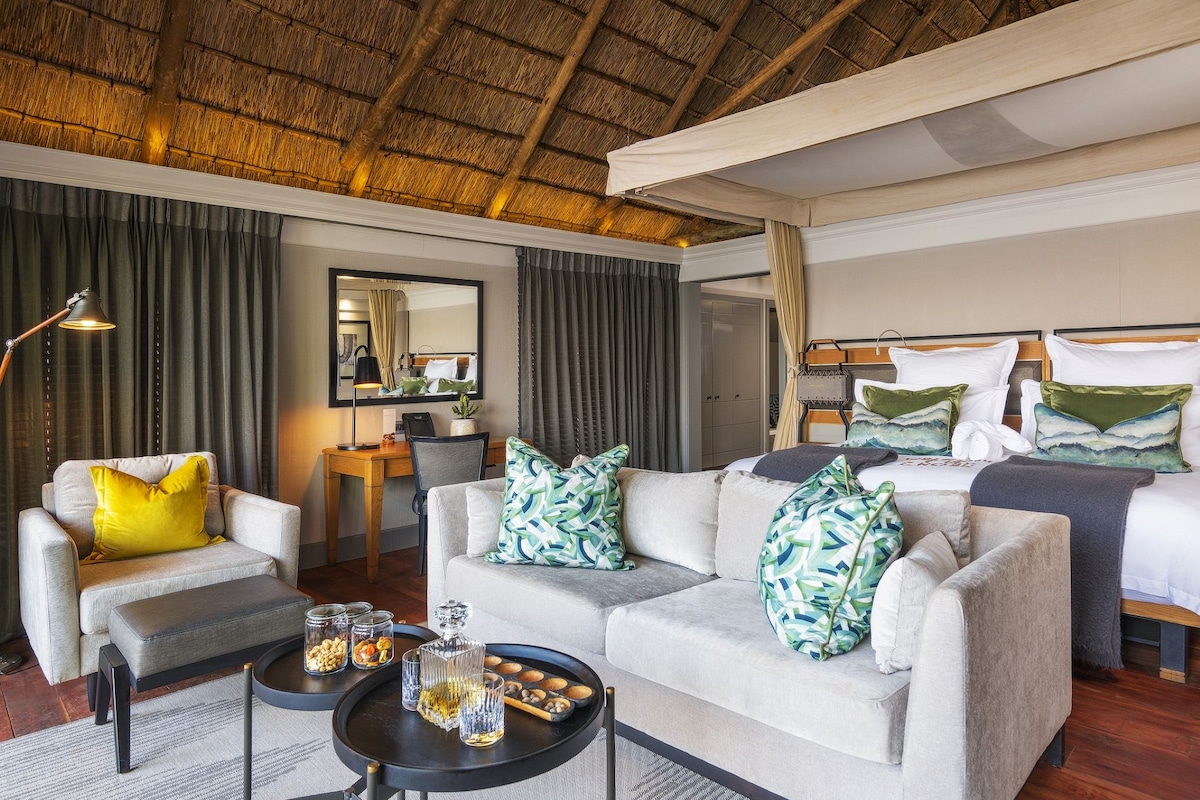 A hotel room with a thatched roof, a sofa set, a bed with pillows, a desk with a lamp, and a mirror.