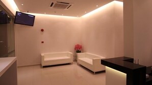 Una moderna zona de recepción con un televisor de pantalla plana montado en la pared, dos sofás blancos, un mostrador de recepción negro y un jarrón con flores rosadas.