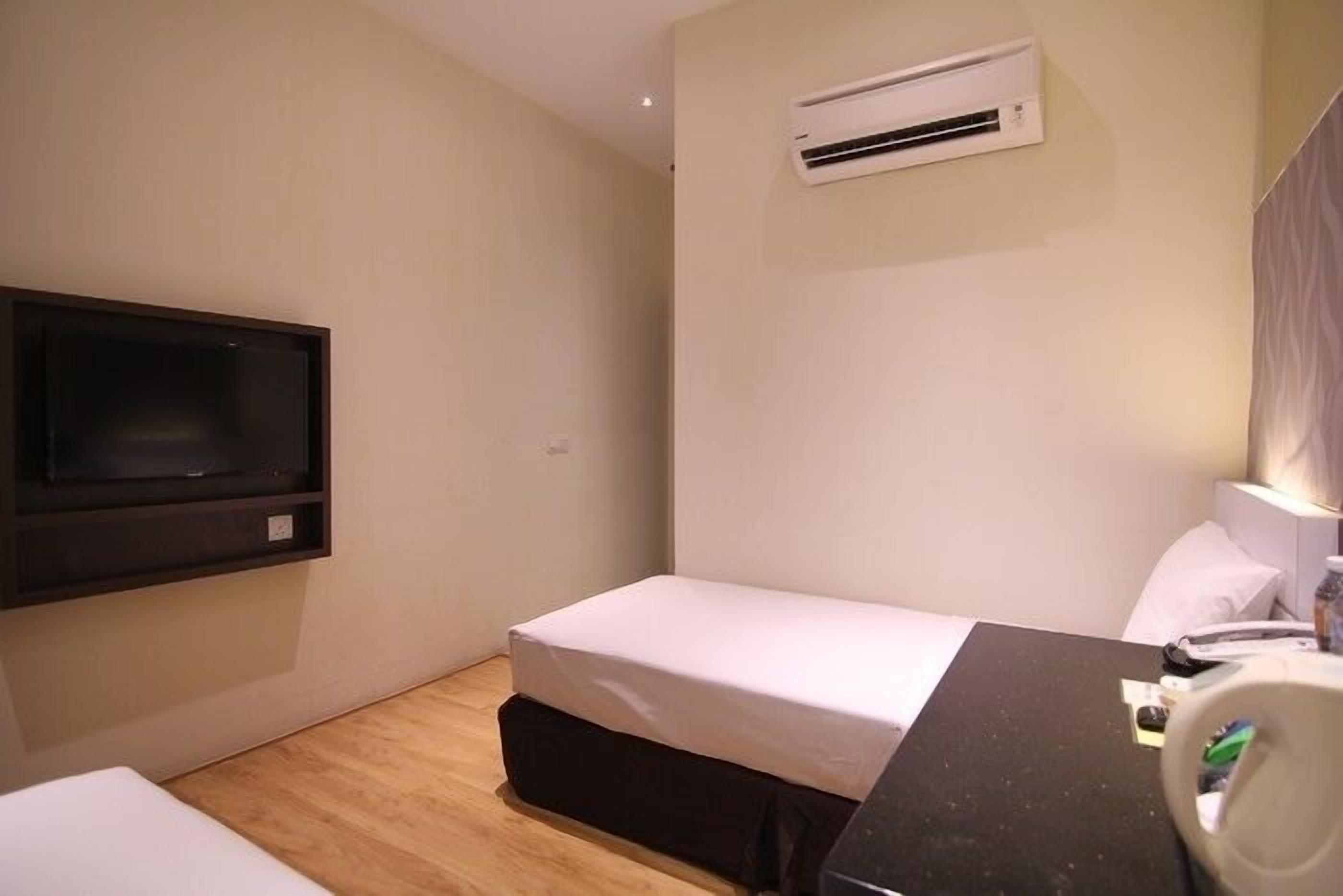 A small, modern hotel room with a single bed, a desk with a chair, a flat-screen TV mounted on the wall, and an air conditioning unit.