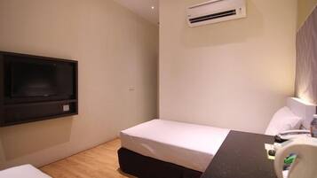 A small, modern hotel room with a single bed, a desk with a chair, a flat-screen TV mounted on the wall, and an air conditioning unit.