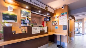 A reception desk with a 'Bienvenue chez B&B' sign, a digital screen displaying hotel information, and a 'DAE' directional sign.