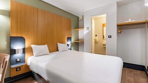 A hotel room with a large bed, wooden headboard, bedside tables with lamps, a wooden floor, and a door leading to a bathroom.