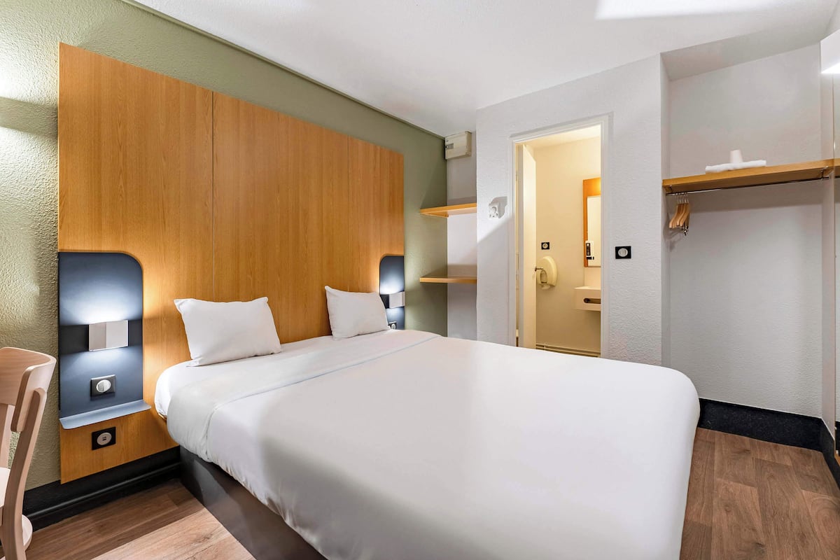 Habitación de hotel con una cama grande, cabecera de madera, mesitas de noche con lámparas, piso de madera y una puerta que da a un baño.