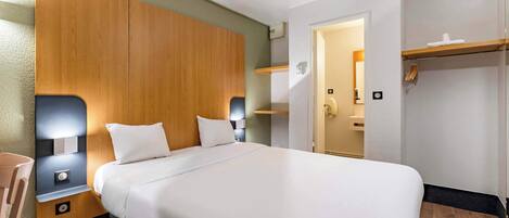 A hotel room with a large bed, wooden headboard, bedside tables with lamps, a wooden floor, and a door leading to a bathroom.
