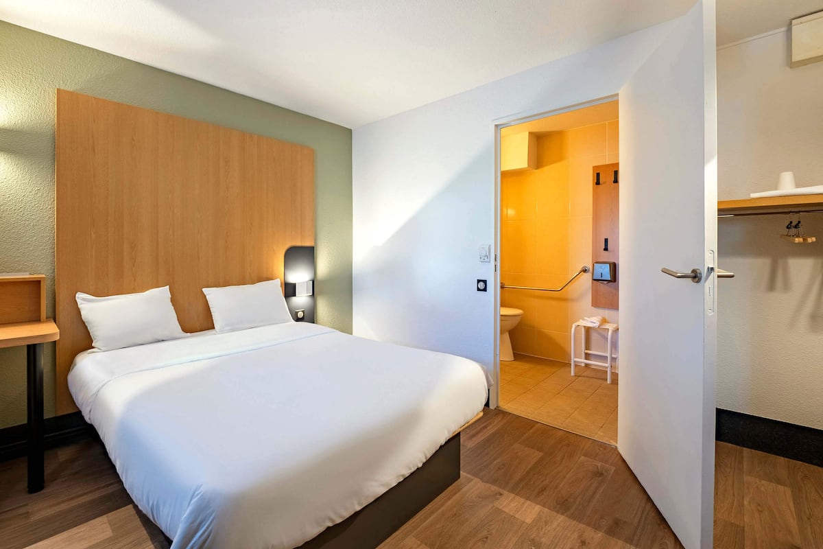 Habitación de hotel con una cama grande, cabecera de madera, mesita de noche y baño visible a través de una puerta abierta.