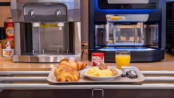 Una mesa con un desayuno que incluye un croissant, huevos revueltos, jugo de naranja, una máquina de café y un dispensador de jugo.