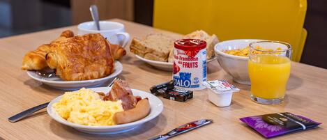 A breakfast table with croissants, scrambled eggs, bacon, cereal, juice, and a can of yogurt.
