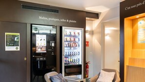 Un hall d’hôtel avec un distributeur automatique, deux fauteuils, une petite table et un extincteur.