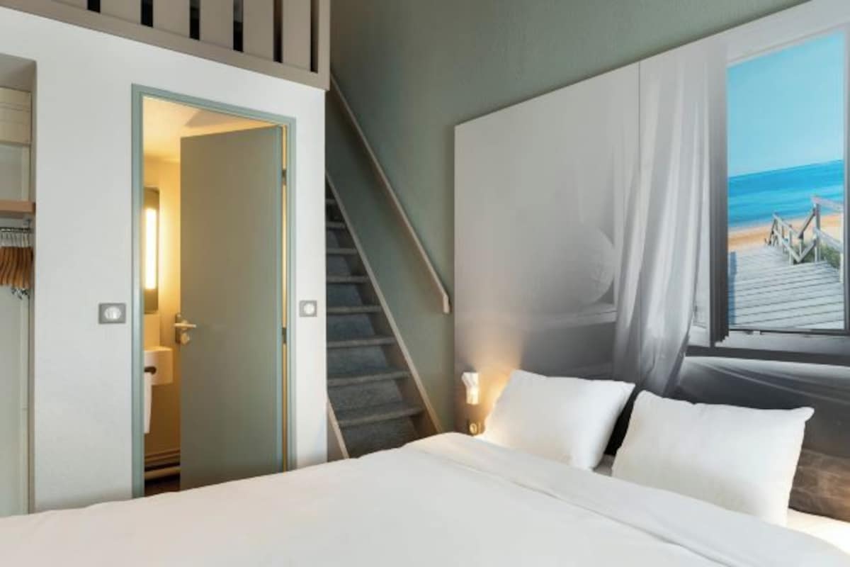 A hotel room with a bed, a staircase, and a beach view through a window.
