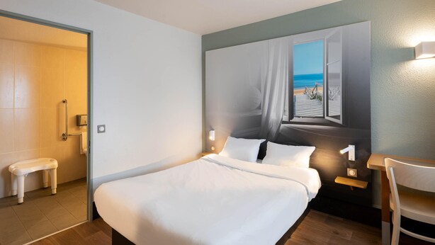 Une chambre d’hôtel comprenant un lit, une chaise, une petite table, un banc, un tableau représentant une scène de plage et une porte donnant accès à une salle de bain.