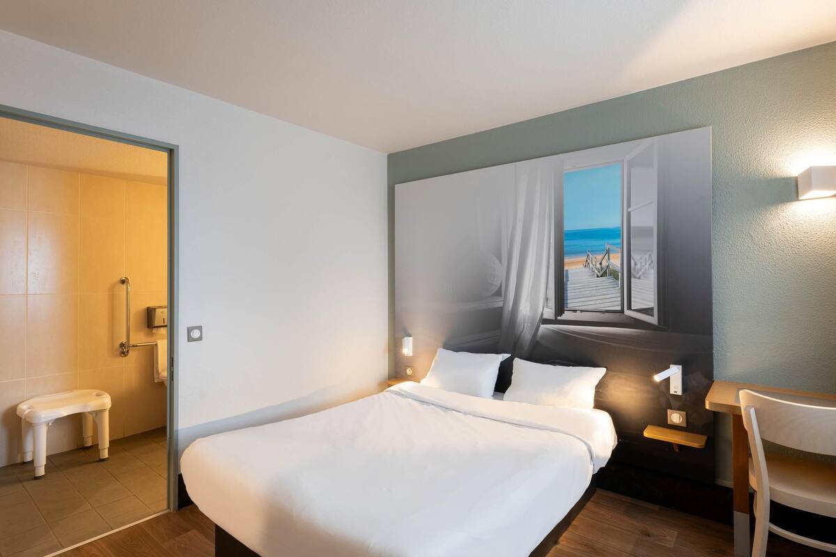 Une chambre d’hôtel comprenant un lit, une chaise, une petite table, un banc, un tableau représentant une scène de plage et une porte donnant accès à une salle de bain.