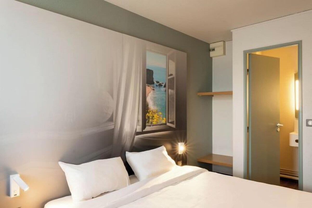A hotel room with a bed, a nightstand, a painting of a beach scene, and a door leading to a bathroom.